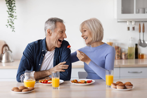 Happy beautiful senior woman having healthy lunch with her husband at home, elderly spouses sitting at kitchen table, eating vegetables and having conversation, feeding each other, copy space - Foto, afbeelding