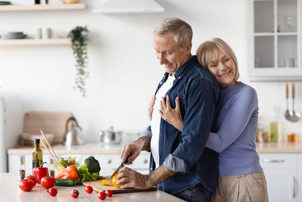 Happy wife attractive senior lady hugging her husband chef cooking healthy delicious dinner for family, loving man taking care of his partner, side view, copy space, white kitchen interior - Photo, image