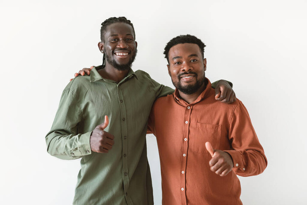 Two Cheerful Black Men Gesturing Thumbs Up Smiling To Camera Approving Offer Embracing Standing Over White Background. Studio Shot Of Guys Showing Like Gesture - Fotó, kép
