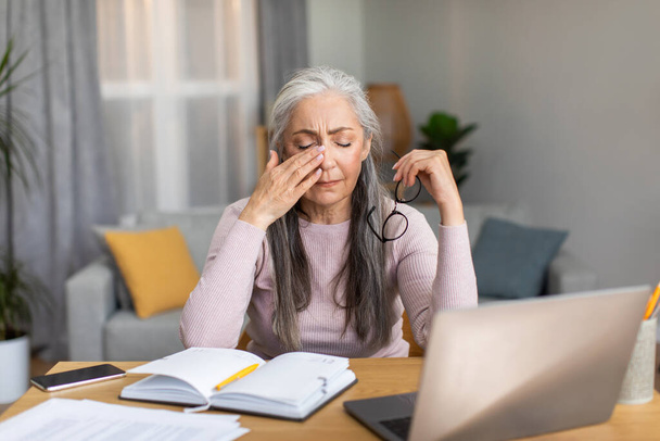 Sad tired caucasian elderly gray-haired woman takes off her glasses, wipes eyes in living room interior. Overwork, vision problems, work at home, business with technology during covid-19 quarantine - Foto, Imagen