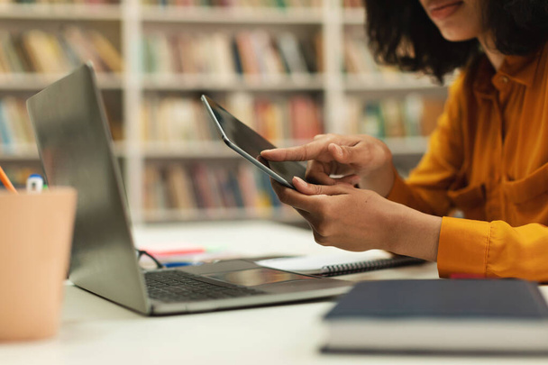 Unrecognizable female student using tablet and laptop for online studies, sitting in library. Latin lady taking part in lecture, talking to teacher on web - 写真・画像