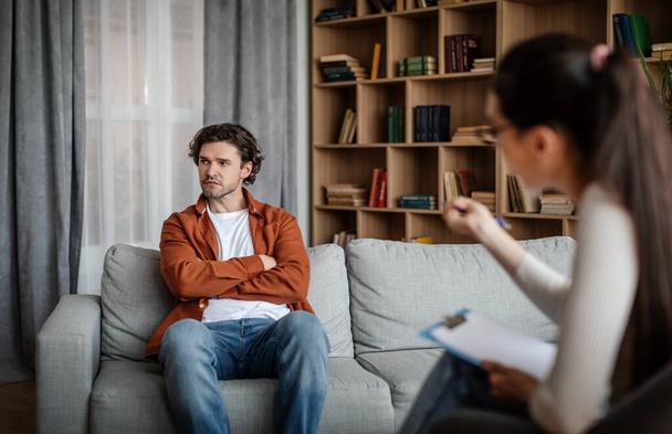 European young woman psychologist talk with dissatisfied guy, patient ignoring doctor advice in clinic interior. Mental problems, professional medical care, fight against depression, help and support - Foto, Imagem