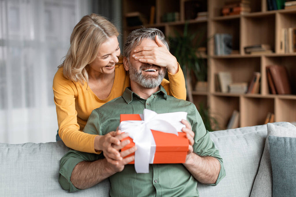 Loving Wife Surprising Middle Aged Husband With Present, Covering His Eyes And Giving Gift Box, Caring Woman Greeting Spouse With Birthday While They Resting Together In Living Room At Home - 写真・画像