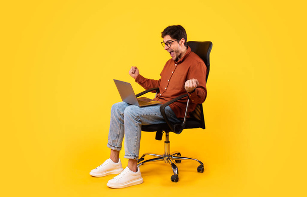 Side View Of Joyful Man Using Laptop And Shaking Fists In Joy Shouting Sitting In Office Chair On Yellow Studio Background. Guy Gesturing Yes Working Online Celebrating Business Success - Φωτογραφία, εικόνα