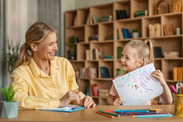 Cute little girl showing attractive woman psychologist her drawing, kid having therapy session with child development specialist at clinic - Foto, Imagen