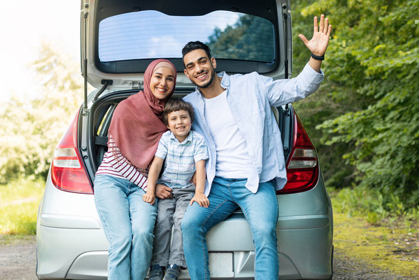 Family travel at own new auto. Satisfied millennial islamic male and female in hijab with son, sitting in car trunk and waving hand, resting from trip together. Vacation, rest from trip and emotions - Foto, Bild