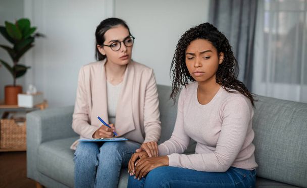 Mental medical support. Sad young european lady doctor in glasses calms black woman patient in office clinic interior. Session with professional, help from depression, psychotherapy and health care - Fotografie, Obrázek