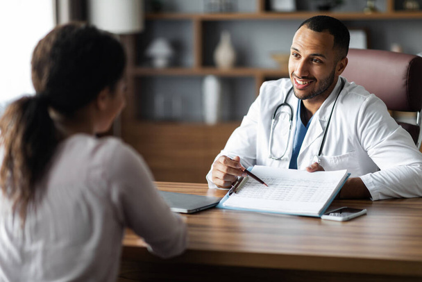 Friendly handsome african american young man doctor having conversation with female patient, practitioner showing treatment plan and smiling, giving details, modern clinic interior - 写真・画像