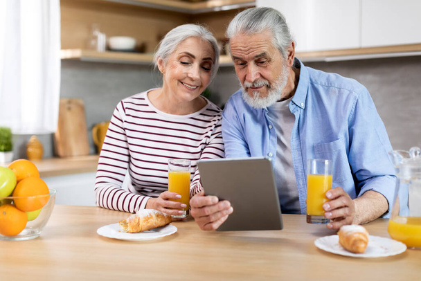 Married Elderly Couple Using Digital Tablet While Having Breakfast In Kitchen Together, Happy Senior Spouses Shopping Online Or Watching Videos While Eating Snacks At Home, Closeup Shot - Фото, изображение