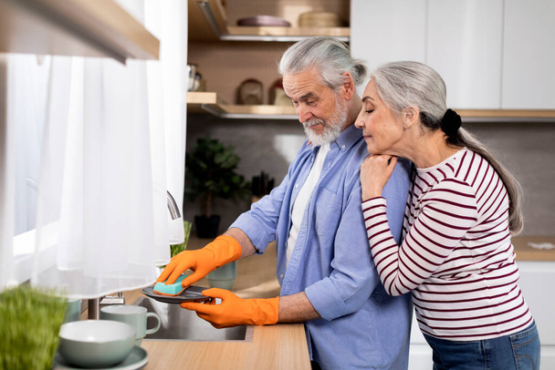 Loving Senior Woman Embracing Her Husband From The Back While He Washing Dishes In Kitchen, Happy Elderly Spouses Sharing Domestic Chores, Caring Husband Helping With Cleaning, Side View - Фото, изображение