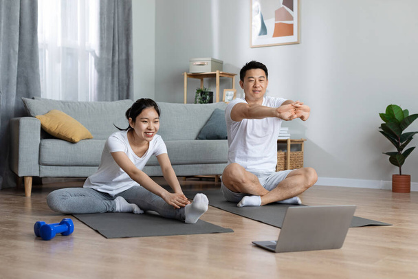 Cheerful japanese man and woman in sportswear sitting on yoga mats in front of laptop, looking at computer screen and exercising, enjoying online fitness video, home interior, copy space - Foto, afbeelding