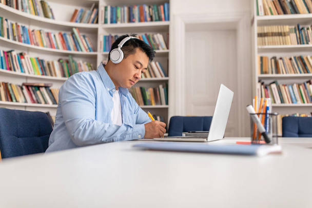 Side View Of Korean Man Working Or Learning Online On Laptop Taking Notes Wearing Headphones Sitting At Desk In Study Room With Bookshelves. E-Learning, Freelance Career Concept - Fotó, kép
