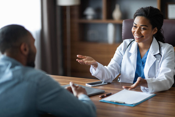 Friendly attractive african american young woman doctor having conversation with male patient, general practitioner consulting young man, gesturing and smiling, giving details, modern clinic interior - Zdjęcie, obraz