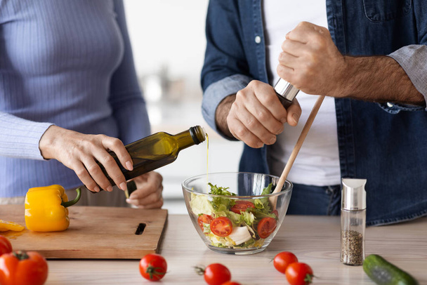 Unrecognizable elderly man and woman in casual outfits cooking together at home, making delicious vegetable salad, add olive oil and seasoning, kitchen interior. Healthy diet for seniors - Photo, image