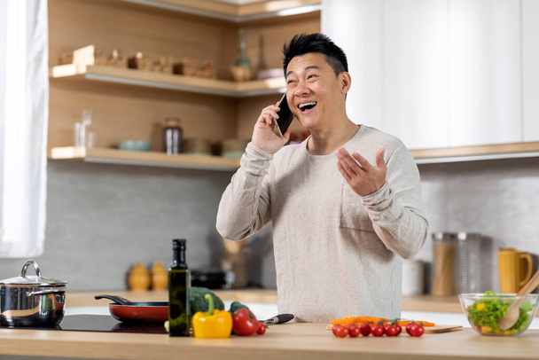 Happy middle aged chinese man in casual outfit standing next to kitchen table at home, cooking healthy meal and talking on mobile phone, laughing, gesturing and looking at copy space - Foto, immagini