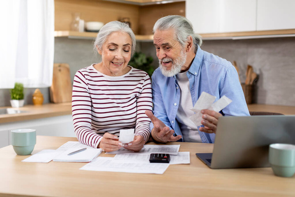 Smiling Elderly Spouses Checking Receipts While Planning Family Budget At Home, Happy Married Senior Couple Counting Monthly Spends, Sitting At Table With Laptop In Kitchen, Closeup Shot - 写真・画像