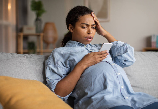 Doomscrolling. Shocked Pregnant African American Lady Using Cellphone Reading Negative News Browsing Internet Sitting On Couch At Home. Pregnancy Lifestyle Concept - Foto, Imagen