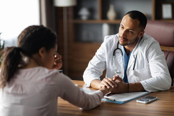 Middle eastern man doctor consoling upset crying sick african american woman patient, therapist supporting sad client, holding hand, giving recommendations, clinic interior. Doctor etiquette concept - Foto, Bild