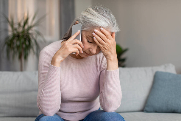 Depressed unhappy caucasian elderly gray-haired lady speaks by phone and holds her head with hand in living room interior. Problems, bad news, stress, pressure, technology during covid-19 quarantine - 写真・画像