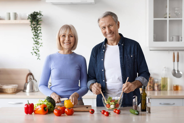 Photo of cheerful senior man and woman cooking together at home, making delicious vegetable salad, smiling at camera, kitchen interior, copy space. Healthy diet for elderly people - Photo, image