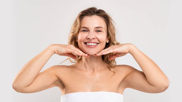 Skincare For Aging Skin. Portrait Of Beautiful Smiling Middle Aged Woman Touching Face With Hands And Looking At Camera, Attractive Mature Female Standing Wrapped In Towel On Light Studio Background - Zdjęcie, obraz