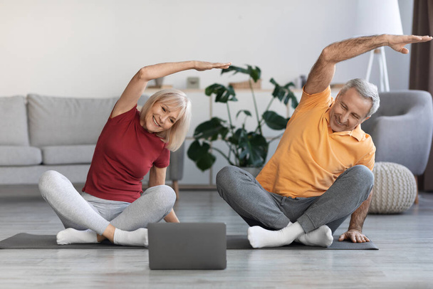 Sporty senior spouses training at home, smiling mature man and woman having workout together, using modern laptop, watching fitness class on Internet, home interior, copy space - Foto, afbeelding