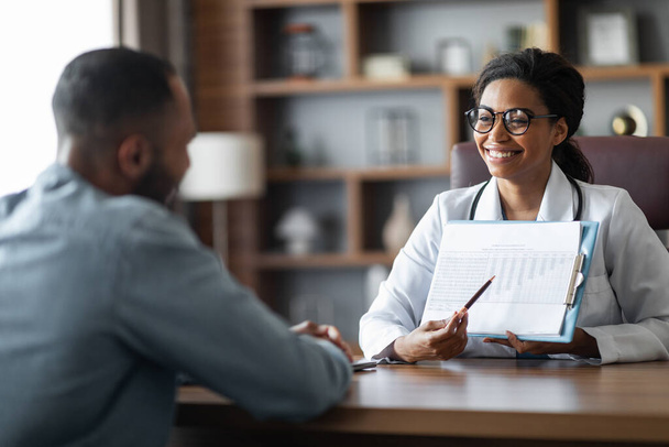 African american young man visiting female doctor at modern clinic, pretty young black lady practitioner holding medical chart, having conversation with guy patient, copy space - Fotoğraf, Görsel