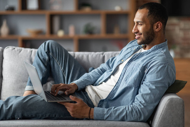 Smiling Young African American Man Using Laptop While Relaxing On Couch At Home, Millennial Black Guy Browsing Internet On Computer, Shopping Online Or Messaging In Social Media, Side View - Photo, image