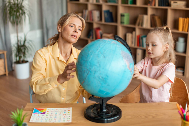 Professional preschool teacher teaching cute girl about globe, having private home class, sitting at desk in living room. Pretty focused kid looking at map with interest - Zdjęcie, obraz
