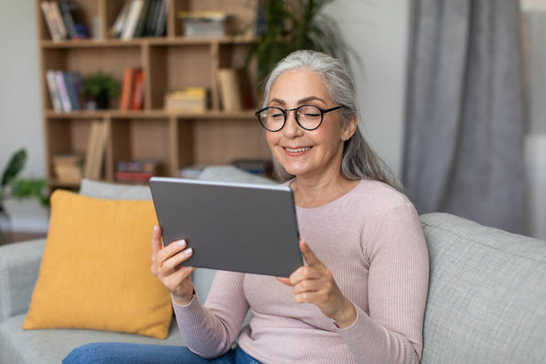 Happy caucasian aged gray-haired lady in glasses chatting on tablet, watch video lesson in living room interior. Modern tech, work remotely, meeting and video call at home during covid-19 pandemic - Foto, imagen