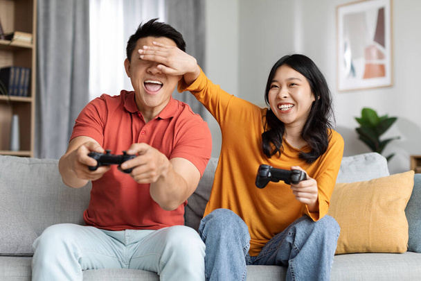 Cute chinese lovers having fun at home, happy asian man and woman sitting on couch, holding joysticks, playing video games, laughing lady covering her boyfriend eyes, cheating in game - Foto, Imagen