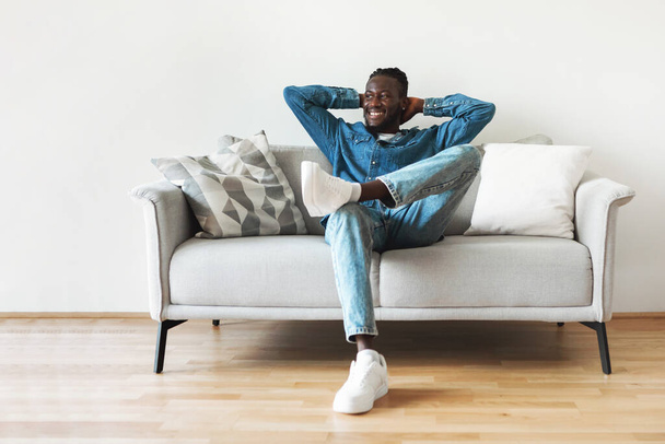 Щасливий чорний хлопець сидить на дивані тримає руки за голову розслабляючись, дивлячись в сторону насолоджуючись вихідні вдома. Тисячолітній чоловік відпочиває у сучасній вітальні
 - Фото, зображення