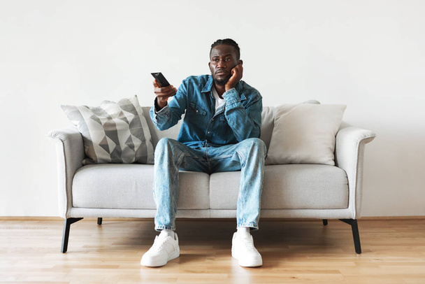 Bored Black Guy Watching Boring Movie On TV Holding Remote Controller Sitting On Couch At Home On Weekend. Television Programming Problems, Bad Film Concept. Front View - Foto, imagen