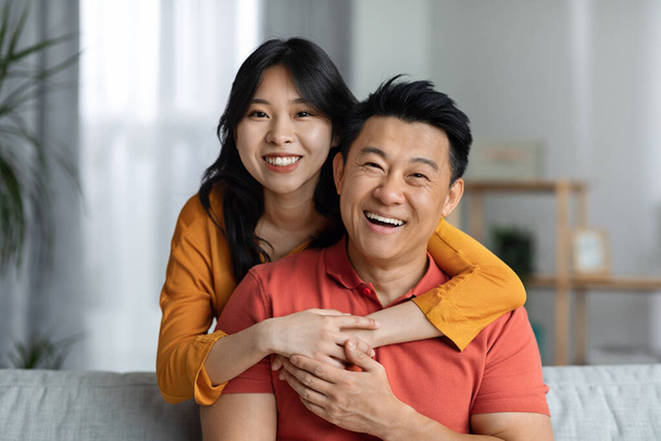 Happy asian man and woman in casual outfits sitting on couch in living room, hugging and smiling, posing together, enjoying weekend at home, having fun, copy space, closeup portrait - Foto, Imagem