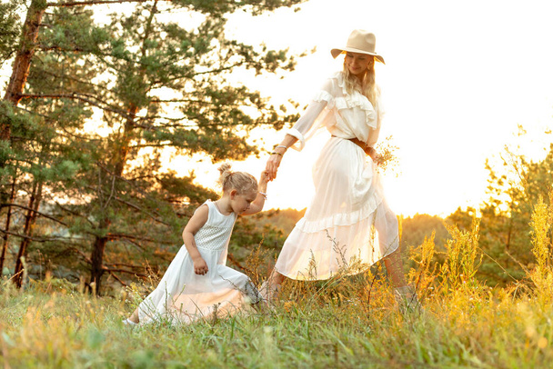 Portrait of happy family wearing white dresses, walking on tall grass among trees in park forest illumined by sunset in summer. Young woman mother holding hand of little girl daughter. Relationship. - Photo, image