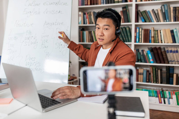 Chinese Male Tutor Having Online Lecture Via Video Call On Phone Pointing At Whiteboard Sitting At Desk In Library Indoors. E-Teaching Concept. Selective Focus On Teacher - Photo, Image