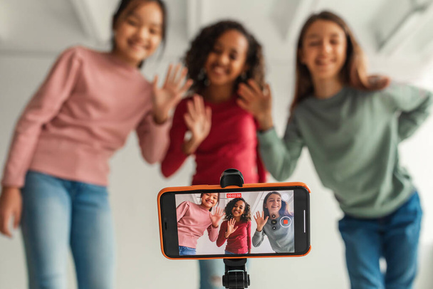 Hello. Three Multicultural Girls Waving Hands To Mobile Phone Making Video For Blog Posing Indoors. Children And Blogging Concept. Selective Focus On Cellphone Screen - Fotografie, Obrázek