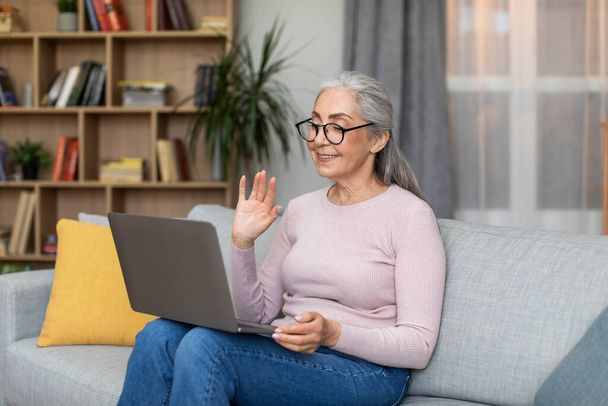 Smiling caucasian senior gray-haired female in glasses waving her hand at laptop webcam in living room interior. Modern device, chat in social networks, meeting remotely and communication due covid-19 - Foto, Imagem