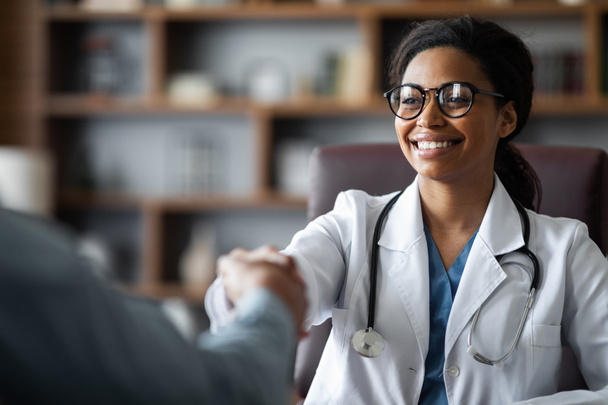 Closeup of friendly young african american lady doctor smiling and shaking male patient hand, cheerful black woman therapist greeting visitor at her clinic, outstretching hand, copy space - Φωτογραφία, εικόνα