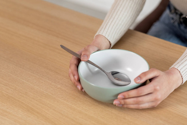 Cropped of woman sitting at wooden kitchen table, holding hands on empty plate bowl with spoon in it, unrecognizable poor hungry starving lady asking for food, panorama with copy space - Фото, изображение