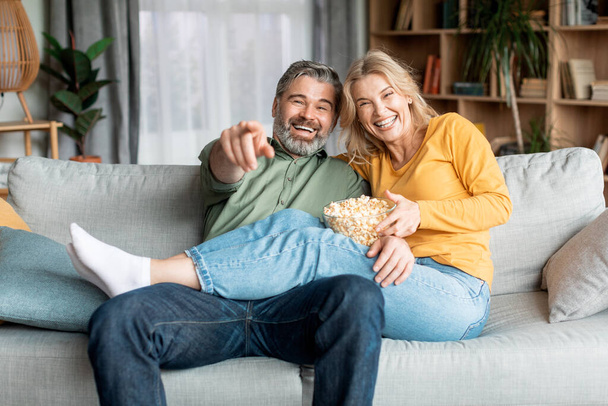 Positive middle aged spouses watching tv and eating popcorn at home, cheerful mature couple having fun while relaxing on couch in living room, laughing together and pointing at camera, closeup - Fotó, kép
