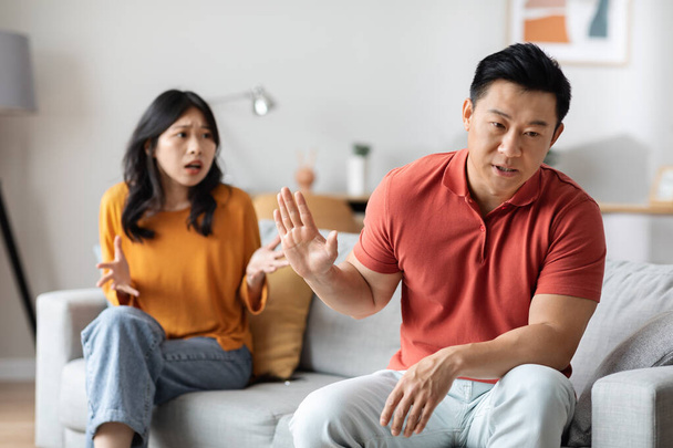Emotional asian couple having fight at home, angry chinese husband and wife sitting on couch, yelling and gesturing, having difficulties in marriage. Divorce, breakup, crisis in relationship - Zdjęcie, obraz
