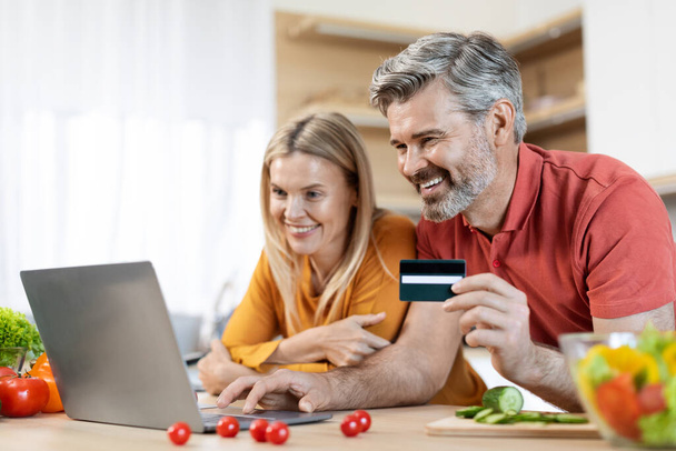 Beautiful couple making online order from supermarket while cooking, standing at kitchen, using modern computer and credit card, purchasing food on Internet, copy space. Easy shopping concept - Zdjęcie, obraz