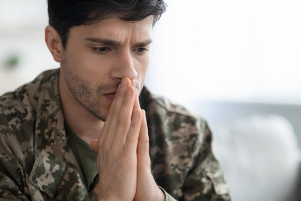 Serious short-haired young man in military uniform soldier looking down, holding palms by mouth, upset guy suffering from posttraumatic syndrome after service, closeup photo, panorama with copy space - Photo, Image