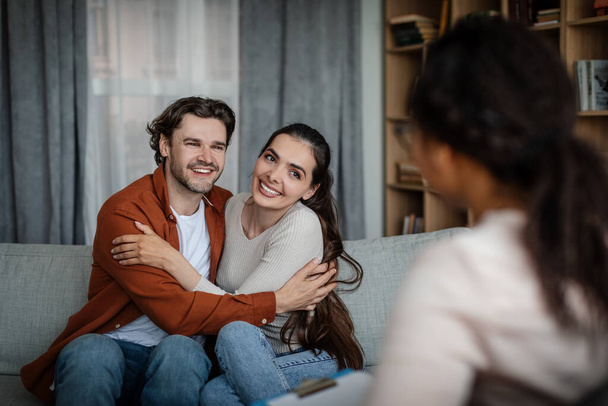 Happy millennial caucasian woman and man hugging on consultation with psychologist in clinic interior. Professional medical care, family relationships, solving mental problems and reconcile couple - Foto, afbeelding