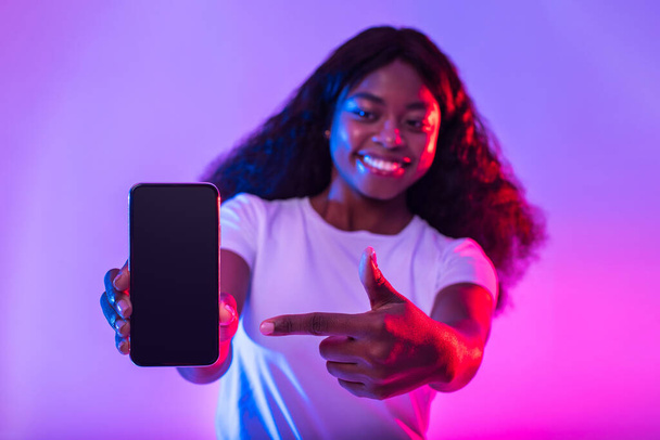 Great mobile offer. Happy young black lady pointing finger at smartphone, advertising new app in neon light, selective focus. African American woman offering mockup for website on cellphone screen - Fotó, kép