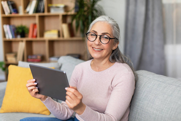 Smiling mature european gray-haired woman in glasses hold tablet, chatting, watch video in living room interior. Social networks, blog, video call and new app, health care during covid-19 pandemic - Foto, Imagem