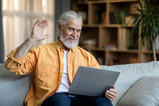 Positive bearded senior man in casual outfit sitting on couch at home, having online party with family, using modern computer, looking at laptop screen, smiling and waving, copy space - Foto, Imagem