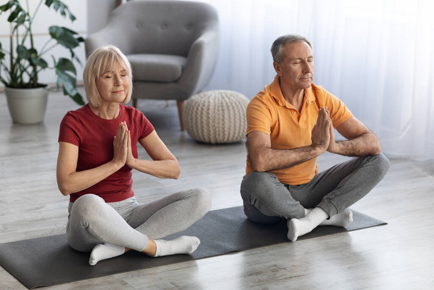 Peaceful senior couple in sportswear sitting on fitness mat in lotus pose with eyes closed, meditating together at home. Elderly people husband and wife having yoga practice, relaxation concept - Foto, Imagen