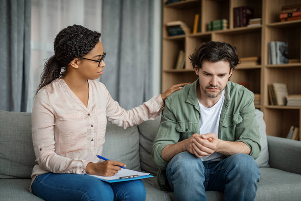 Serious young black female doctor calms european guy, client suffering from depression in office clinic interior. Medicine, psychological therapy, session with professional, mental help and emotions - 写真・画像